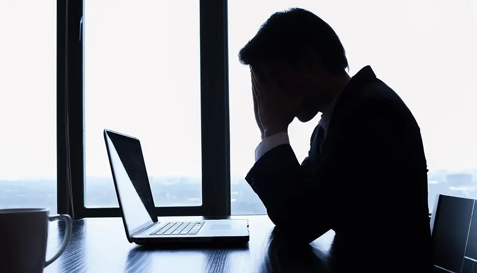 Silhouetted person in a suit, hands on face, seemingly stressed, sitting at a desk with a laptop. Large window in background, creating a tense mood.