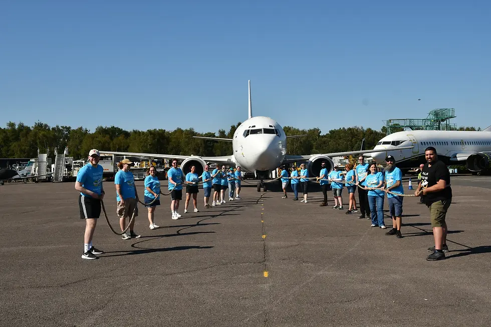 RiskSTOP Group team pull a rope attached to a large airplane on a tarmac. Clear sky, other planes, and trees in the background.