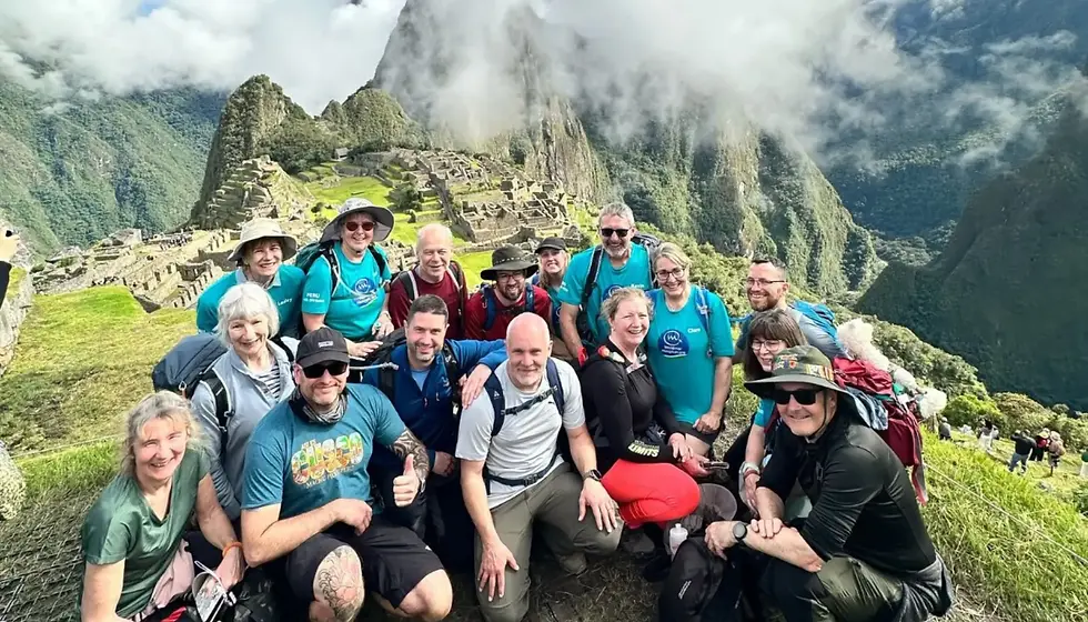 A group of smiling hikers posing at Machu Picchu, wearing colorful shirts and hats. The ancient ruins and stunning mountains are visible in the background.