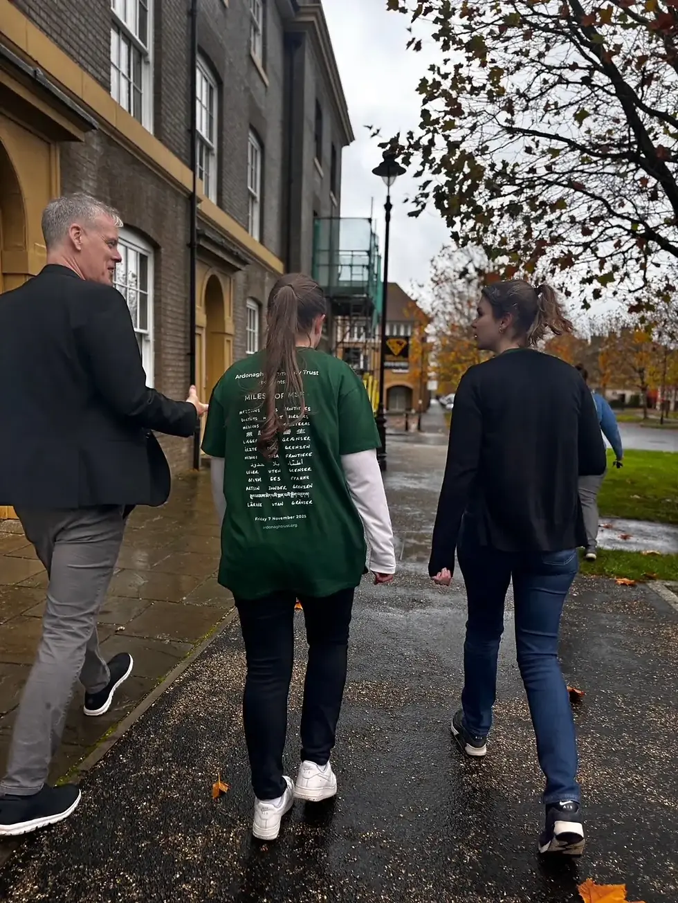 Three people walk through Poundbury, on a wet path beside a brick building. One wears a green shirt with text. Autumn leaves on trees and ground. Overcast day.