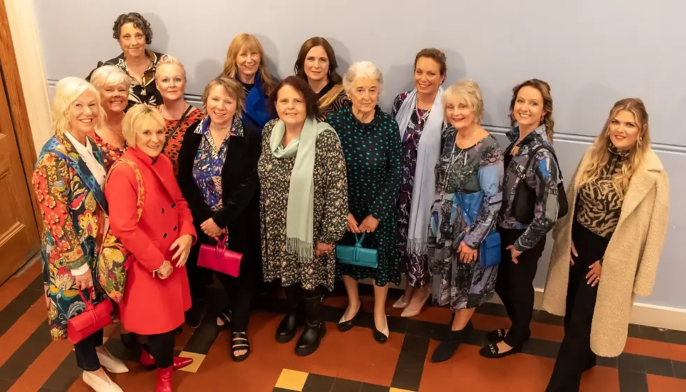 Group of 14 Fashion Show participants smiling in colorful clothing, holding bright purses, standing on patterned floor in a room with a light blue wall.