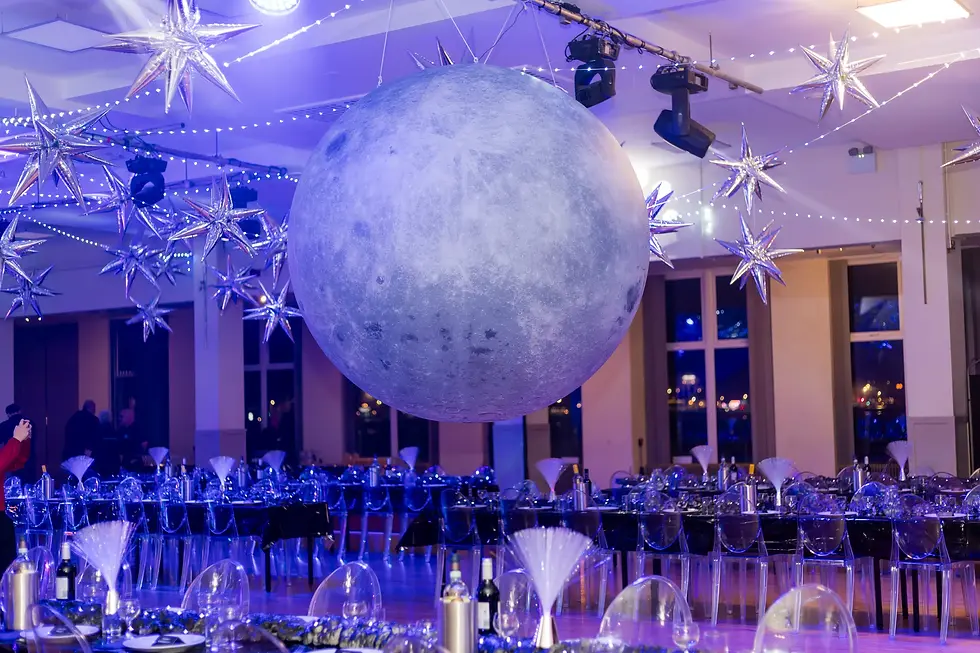 Event room with a giant moon and silver star decorations, blue lighting. Tables set elegantly; festive, cosmic atmosphere.
