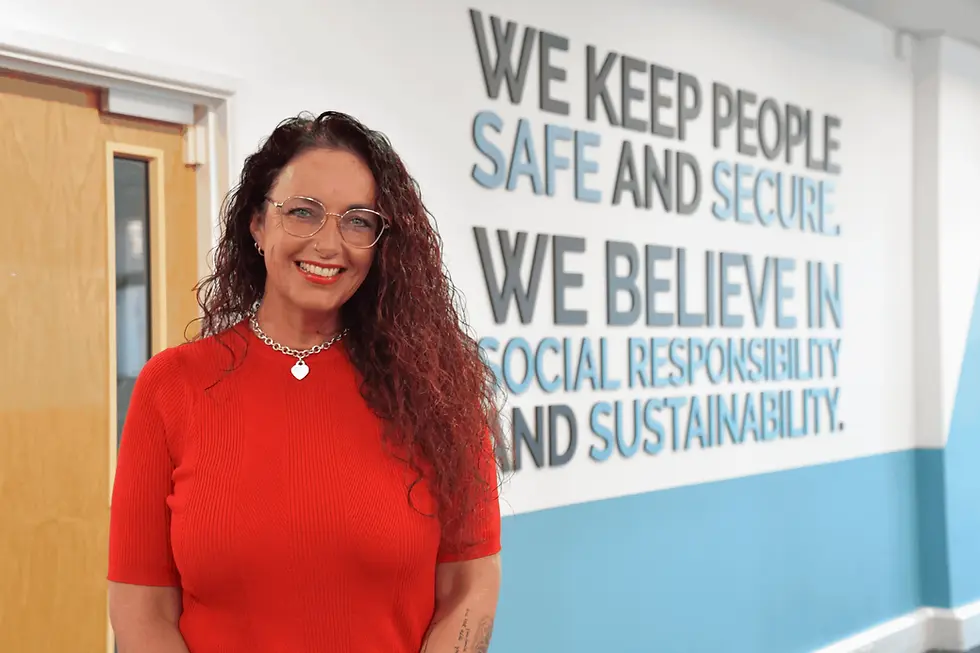Nat Joyce standing and smiling for the camera, stood in the RiskSTOP Group office, in front of the Group's mission wording 'We keep people safe and secure. We believe in social responsibility and sustainability.'