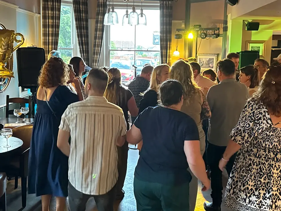 People dancing and socializing in a lively, warmly-lit room with checkered curtains. Balloons and drinks visible, creating a celebratory mood.
