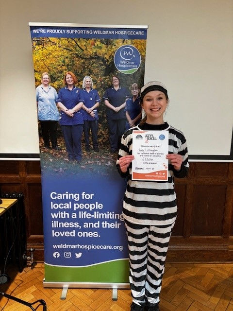 Amy Lillington dressed in prison clothing next to a sign for weldmar hospicecare